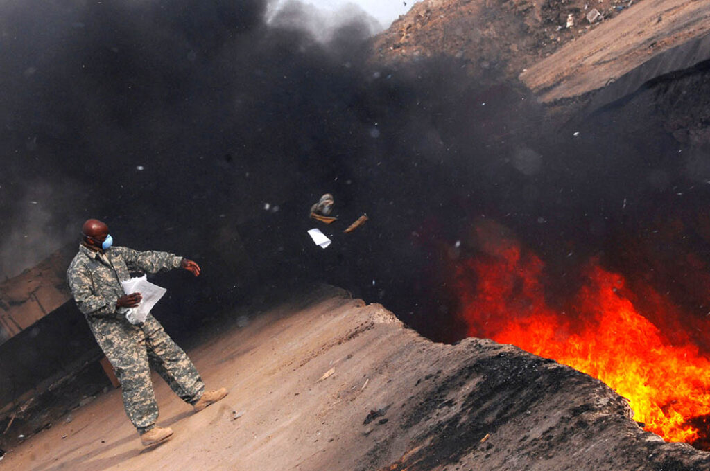 A US soldier tosses unserviceable uniform items into a burn pit on 10 March 2008 at Balad Air Base, Iraq. [USAF/Senior Airman Julianne Showalter]