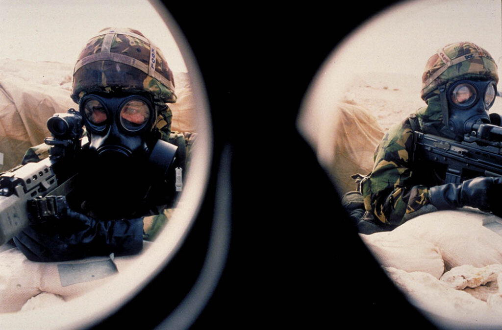 View through the face mask of two British soldiers wearing protective chemical weapons clothing holding rifles in a foxhole during the First Gulf War. Chemical weapons were a major threat during the war and elements believed to be associated with Iraqi capabilities in this area were targeted during some of the air strikes. [Crown Copyright 1991]